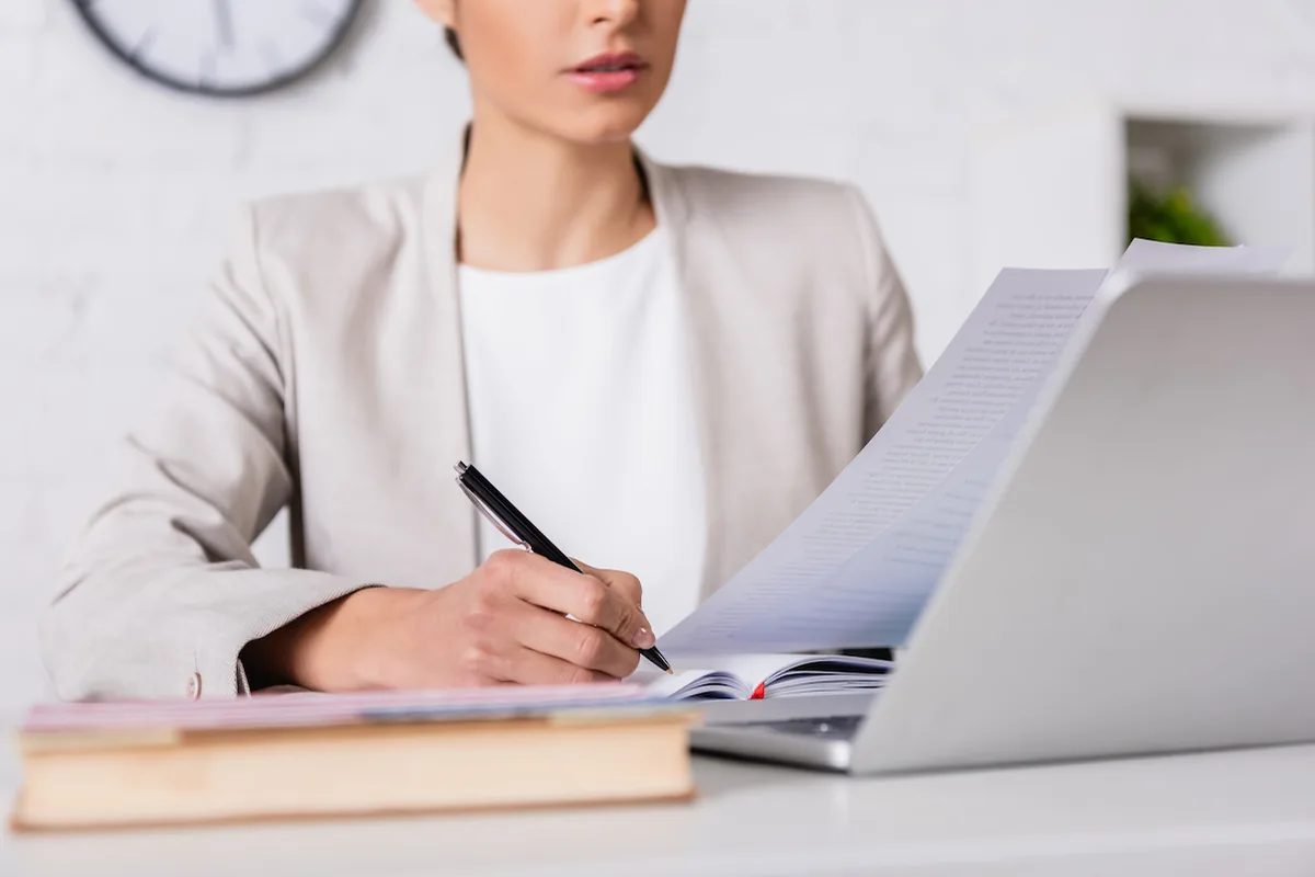 Persona organizando documentos y escribiendo en un cuaderno junto a una computadora.